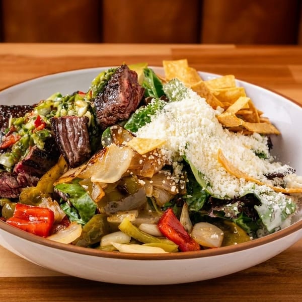 Steak salad with grilled vegetables, cheese, and tortilla strips in a white bowl on a wooden table.