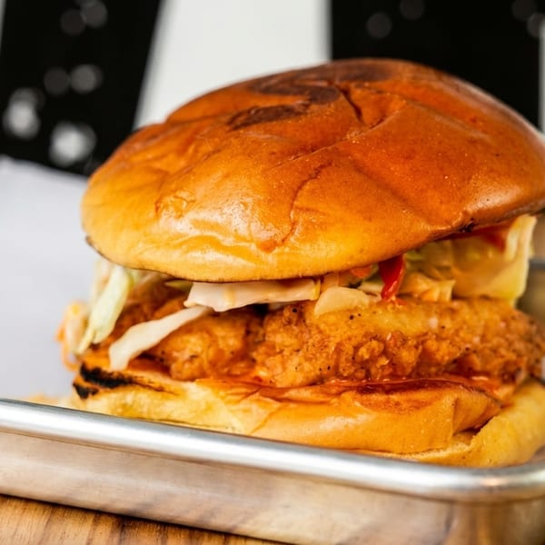 Crispy fried chicken sandwich with slaw and sauce on a brioche bun, served on a metal tray.