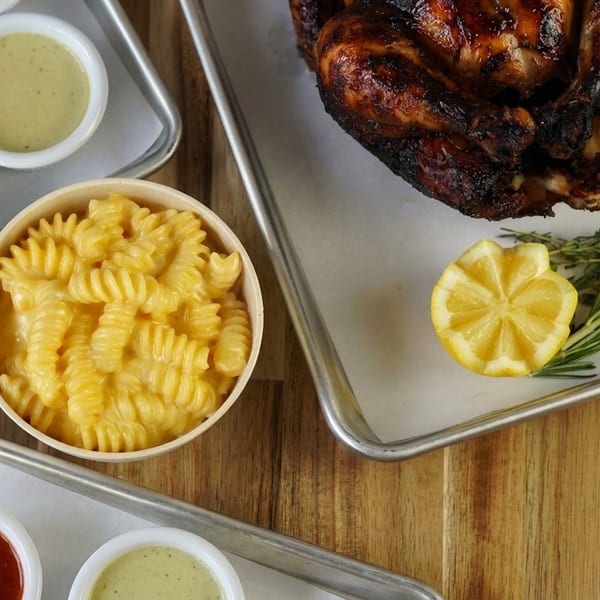 Grilled chicken, mac and cheese, and various dipping sauces on metal trays on a wooden table.